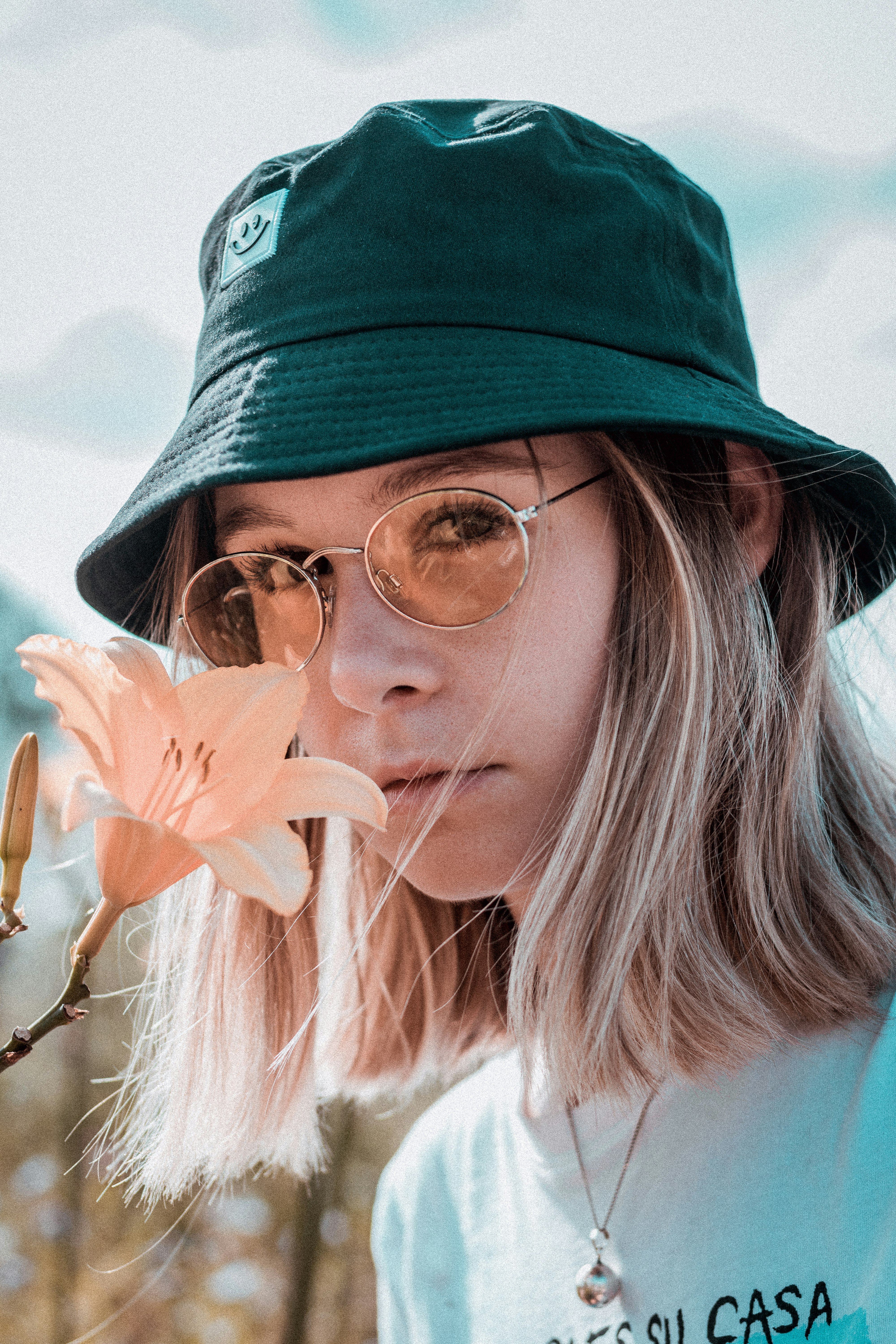 woman wearing white and black printed crew-neck shirt, eyeglasses, and black bucket hat smelling beige petaled flower